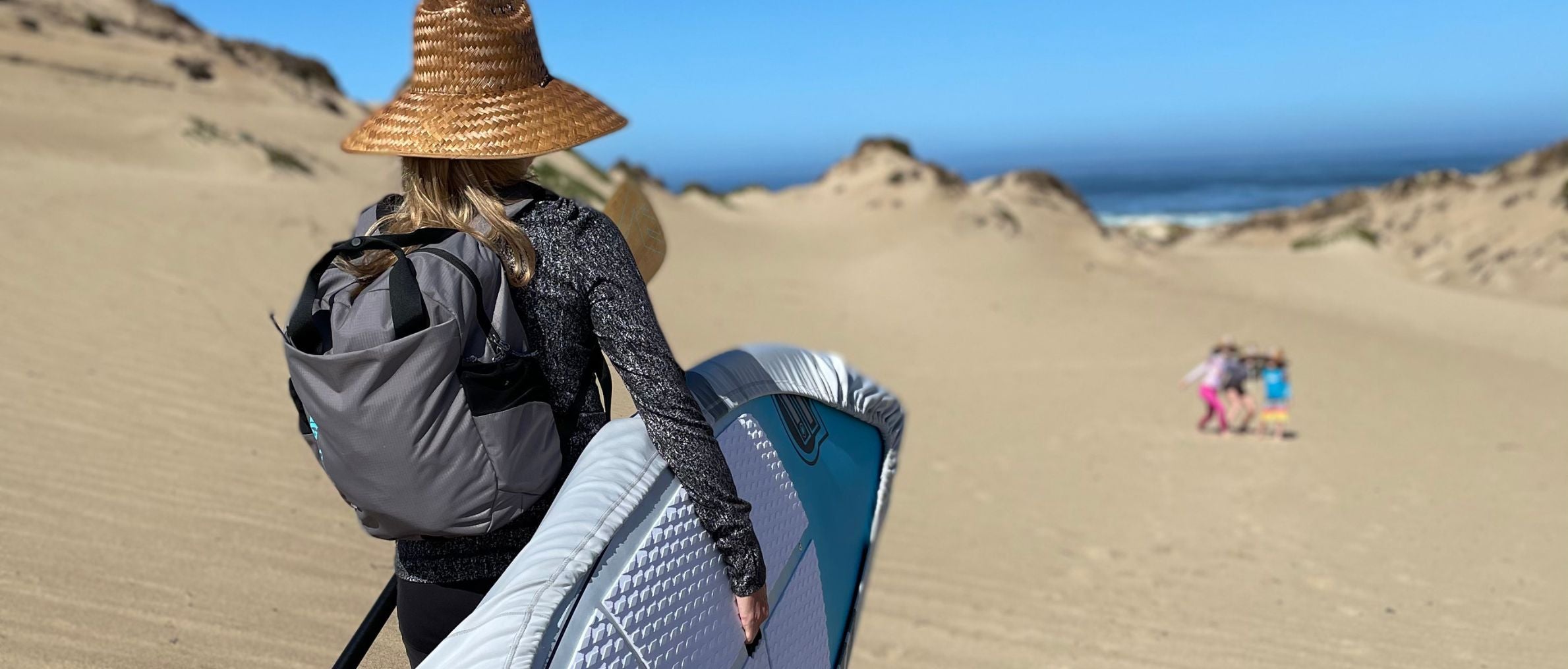 Person with a backpack and hat walking on sand dunes towards the ocean.