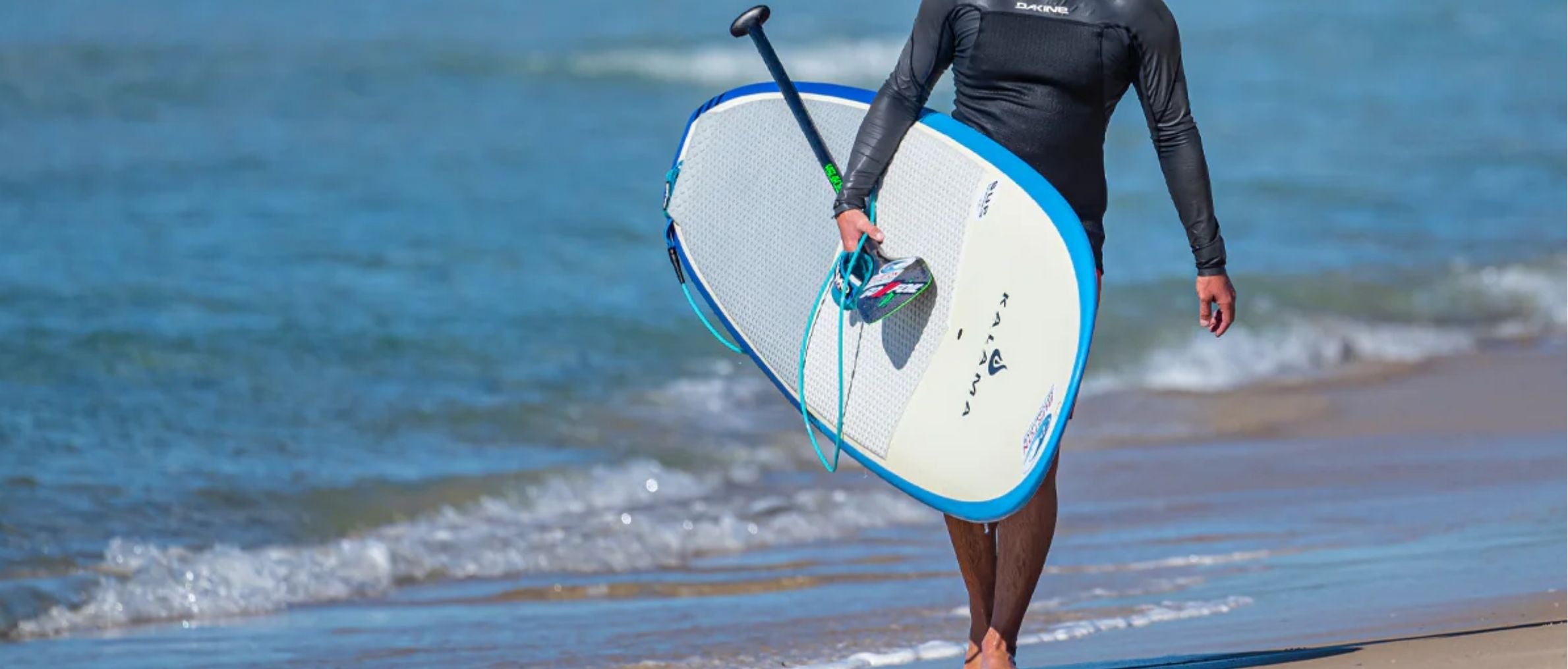 Person in a wetsuit holding a paddleboard with VAMO Surf Leash on a beach with ocean in the background